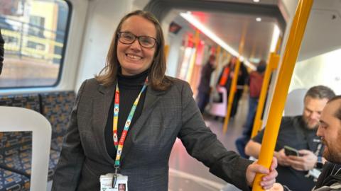 Nexus managing director Cathy Massarella smiling as she hold a yellow hand rail while on board the first new Tyne and Wear Metro in December 2024. She is wearing grey suit jacket over a black turtle neck top. She has glasses and a colorful lanyard. Her brown hair curls at the ends  and reaches her shoulders. Behind her two men are sitting and looking in their phones. Other passengers are standing further back in the train. 