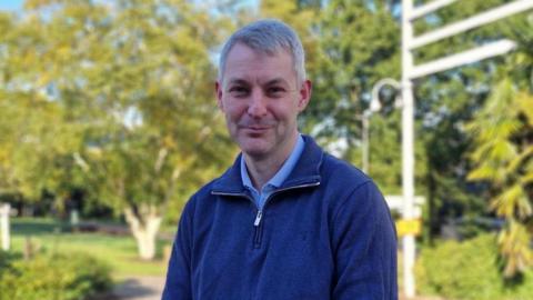 Will Forster looks at the camera and smiles. He is wearing a light blue shirt and a darker blue jumper with a zip neck, behind him there are green trees and a blue sky.