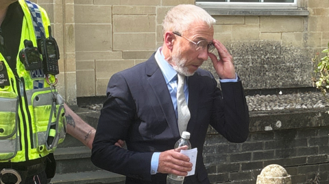 James Holder is excorted from Cirencester Magistrates' Court by a police officer. He is wearing a dark suit and grey tie