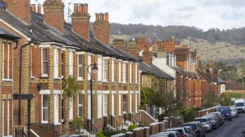A row of redbrick terraced housing. There is countryside in the background and cars are parked along the street. 
