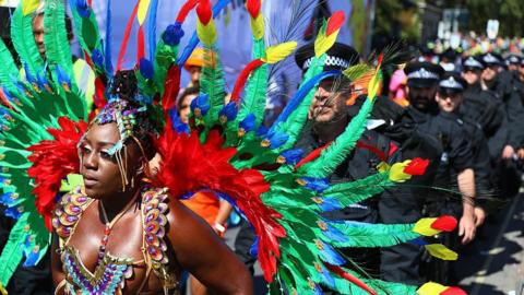 A brightly-feathered parade participant stands in front of a police line during the Notting Hill Carnival on August 25, 2025