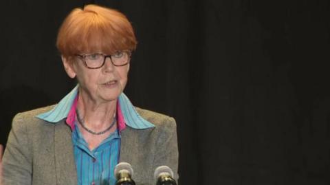 Dame Vera Baird with short ginger hair and wearing glasses, is photographed speaking at a press conference wearing a blue shirt and grey blazer