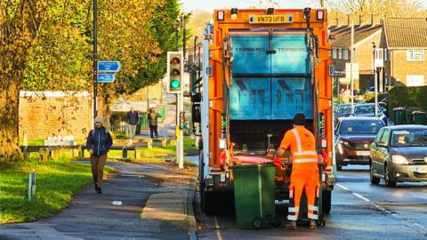 A refuse collection worker, wearing reflective workwear, emptying a rubbish bin into the rubbish truck on a city street.