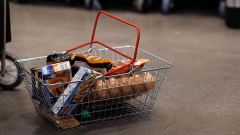 A shopping basket full of beige groceries sits on a grey concrete supermarket floor.