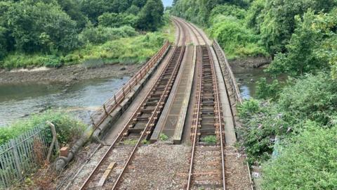 A train line on a bridge. There is a small river under the bridge. Either side of the rail-line there are green trees and bushes. 