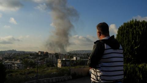 A man in a grey and black striped jacket looks at smoke rising from a building following an Israeli airstrike, he is holding a smart phone. 