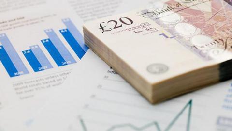 A generic image of bank notes and financial papers. A stack of £20 notes is placed on top of papers with charts and graphs.