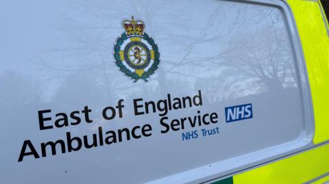 Side of a yellow, green and white ambulance. The words "East of England Ambulance Service NHS Trust" are written on the side of the ambulance, below the trust's logo.