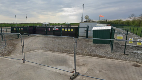 Google shot of the A well site in High Fosham. There are a number of green containers inside a metal perimeter fence and an orange wind sock on a pole.