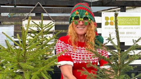 Kim is wearing a red Mother Christmas dress and Christmas tree novelty glasses standing among three Christmas trees