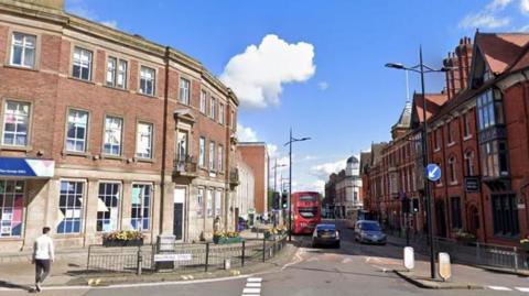 Stafford Street in Wolverhampton city centre with road markings, road signs, a traffic island, lampposts, buildings and shops. There are cars and a bus on the road.