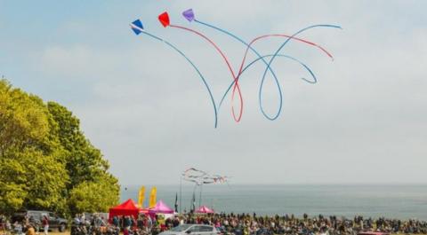 Three coloured kites fly in a display above coloured tents, and an audience of spectators on the cliff above the sea. The sun is out and the sky is blue.