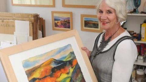 Woman with white/grey hair with an abstract landscape painting in a frame. She is wearing a white top with grey dress