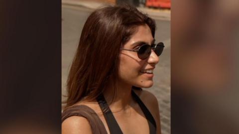 Young woman with long brown hair, wearing sunglasses.