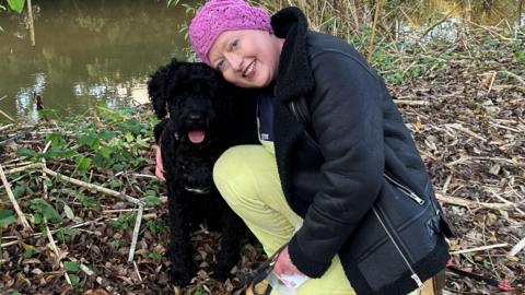 A woman wearing a pink crochet hat, black fur lined jacket and yellow trousers crouches on the ground with her arm around a medium-sized black dog. The dog has curly hair and is sticking its tongue out. Behind the pair is a river.