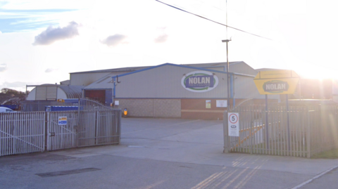 A wide picture of the Nolan Recycling warehouse unit behind open grey gates. The sun is shining in the background of the image.