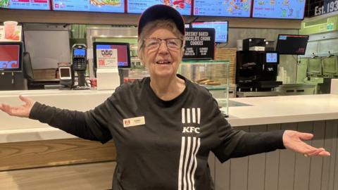 A woman in a black KFC uniform and cap stands in front of a KFC counter. She is smiling and has her arms outstretched