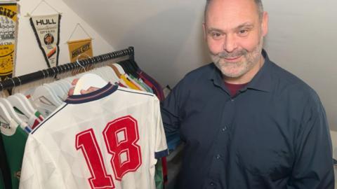 An man with cropped grey hair and stubble holds an original white England shirt from 1988, with the number 18 on the back, on a hanger. There is a rail of shirts behind him and Hull City pennants hanging on the wall. He is wearing a navy coloured short with a burgundy undershirt 