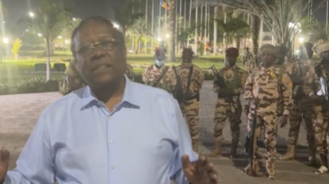 Foreign Minister Abderaman Koulamallah seen in a video apparently shot at the presidential palace in N'Djamena. He is wearing a blue shirt. Behind him are lines of palm trees and a lawn. Over his shoulder is also a group of soldiers in camo uniforms. 