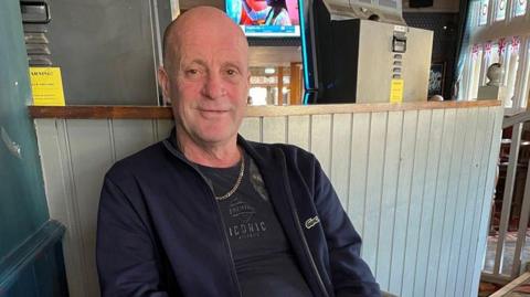 An archive photo of John Smith who is sitting at what appears to be a pub. He is wearing dark clothes and a gold chain around his neck. He is looking at the camera and smiling.