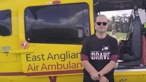 Steve Jones standing in front of a yellow ambulance with East Anglian Air Ambulance on its door. He is wearing dark glasses and a black T-shirt with Brave written on it over a red long-sleeved top, with the sleeves rolled up. 
