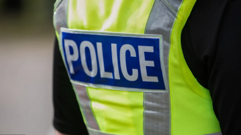 The back view of a British police vest that is illuminous yellow and has the words POLICE in white against blue rectangle background 