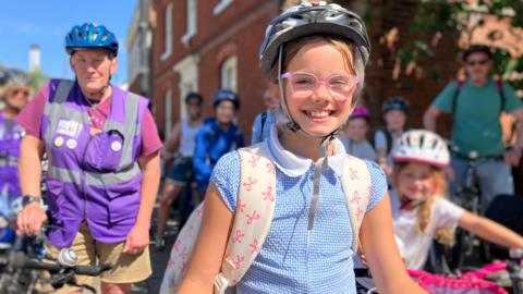 A girl with a bike helmet and school rucksack with others on bikes in the background.