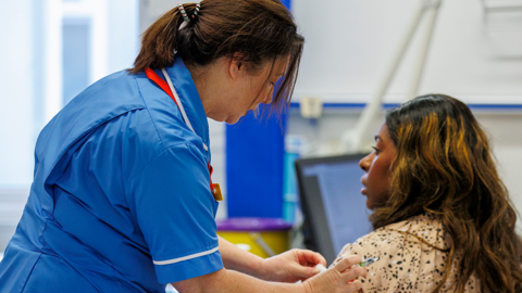 A nurse wearing blue scrubs administers a vaccination to a lady with dark hair and a patterned shirt who is sitting down