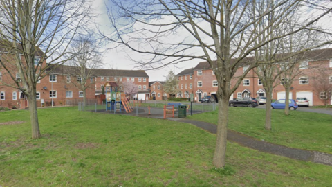 A children's playground pictured in the middle of a housing estate. The playground is surrounded by grass with several trees. There are three storey red brick buildings around the edge of the grass.