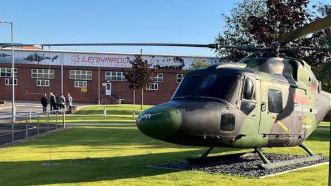 A camo-painted helicopter standing on a green field with a factory building in the background. The word Leonardo is painted across the top of the building.