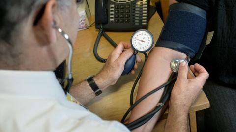 A male doctor wearing a white shirt is photographed from behind while he uses equipment to check a patient's blood pressure.