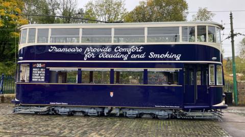 A side-on view of a two-tier navy coloured tram, interspersed with cream coloured lining. Between the top and bottom windows are the words 'tramcars provide comfort for reading and smoking' in a fancy white font.