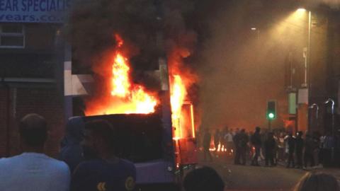 A double-decker bus on fire in Harehills, with flames billowing through smashed windows. People linger on the smoke-filled street nearby. 