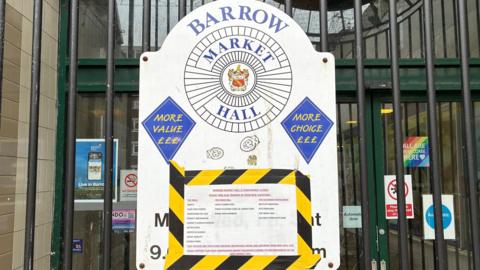 A close-up of the closure sign which is surrounded by yellow and black tape and stuck on the white opening times sign on the black gate. The white sign reads 'Barrow Market Hall' with 'more value' and 'more choice' written on either side. The writing is too small to make out what it says on the closure sign.