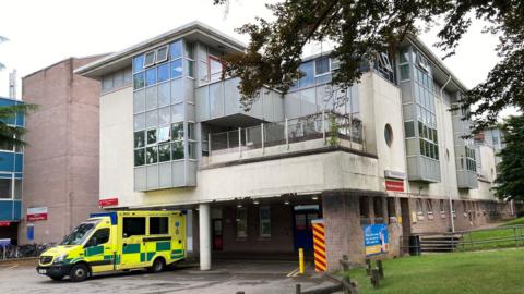 An ambulance waits outside a covered back entrance at an aging hospital, which has marks on its white render. It is about three stories tall with windows jutting out, and there is a green grass verge to the right.