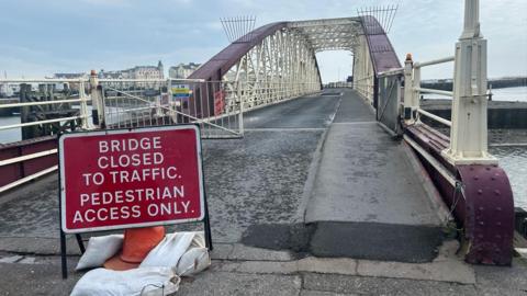 A red road sign that reads 'bridge closed to traffic, pedestrian access only'. It is placed on the road in front of the roadway onto the swing bridge, which is a red and white metal structure.