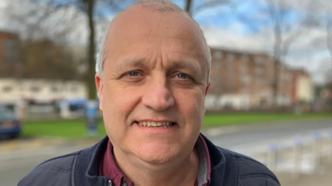 Nick Buckley, with short grey hair and a dark blue jacket, looks straight ahead for the camera