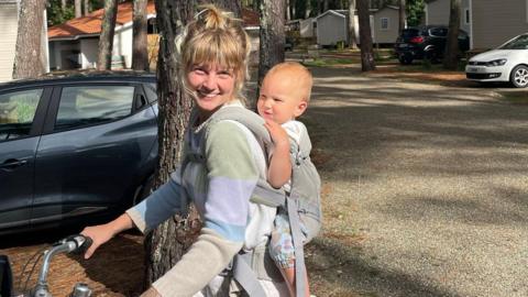 A woman sitting on a bike with a baby in a carrier on her back. The woman is holding the bike handles and is smiling at the camera. They are in a car park. Cars are in parking bays.