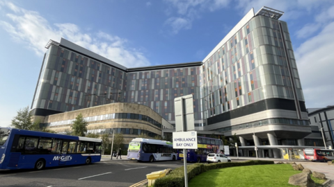 Looking at the many windows of the tall grey hospital building, on a sunny day with blue skies. Several buses and cars are parked in the foreground.