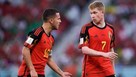 Footballers Hazard and De Bruyne communicating with each other on the pitch. They are both wearing the same red team shirt.