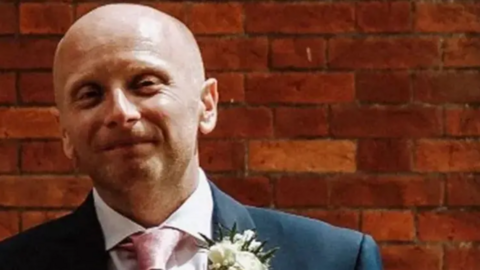 A photograph of Dan Kay, dressed for a wedding, in a blue suit, pink tie, and white buttonhole. 