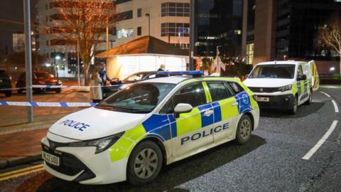 Police cars on a road with blue and white police tape cordon in place following a stabbing. 