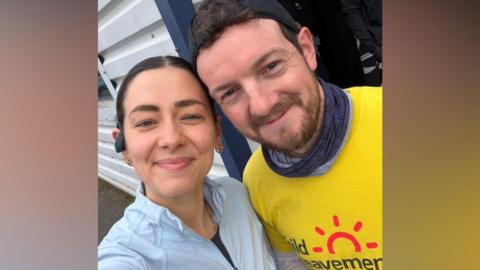 A dark-haired woman wearing a pale blue shirt. She has an earpiece on her right ear. She is standing next to a dark-haired man with a yellow t-shirt, with a red sun logo and the label Child Bereavement printed on the front.