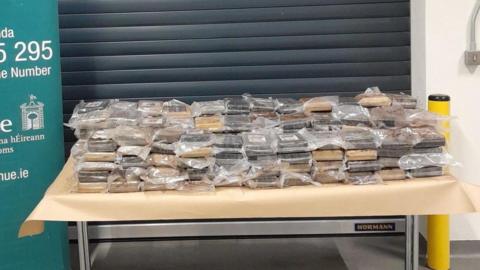 A close-up of a table placed in front of a grey shutter inside a non-descript room. The table has a light brown covering. On top are stacks of brown-looking material in plastic packaging, perhaps two rows with 12 stacks in each row. The drugs on the table are beside a partially-seen sign with the logo and contact details for the Irish Revenue service