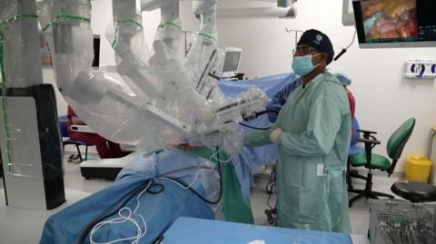 A robot with several arms performing a surgery in an operating theatre. There is a surgeon in a green frock coat who is wearing a face mask. There is a monitor in the background and surgical equipment in the foreground.