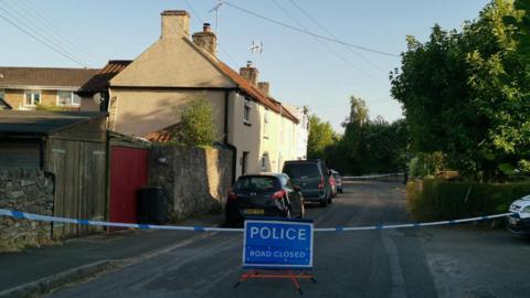 A quiet residential street, with a few houses along one side and trees on the other, is blocked by police tape and a sign which says 'Police, road closed'.