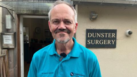 A gp called Dr David Davies stands outside his surgery in Dunster. He has a  short beard and moustache and is wearing a turquoise polo top