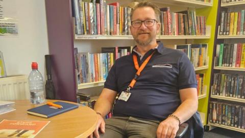 Jimi Wilson wearing a navy polo shirt sits in a chair in a library. He has an orange lanyard around his neck and is setting in front of two bookcases. On the table in front of him is a notepad, a bottle of water with a red cap, a white binder and an orange leaflet.