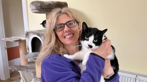 The photo shows a woman smiling at the camera. She has blonde hair and black framed glasses. She's holding a black and white cat, which is also looking at the camera. Behind her, there is a cat tree.