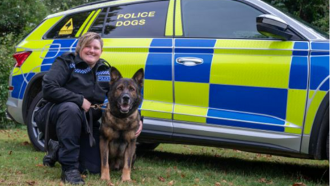 PC Samantha Clark, in her police uniform, smiles as she kneels next to Olly, a German Shepherd, alongside a police vehicle. 